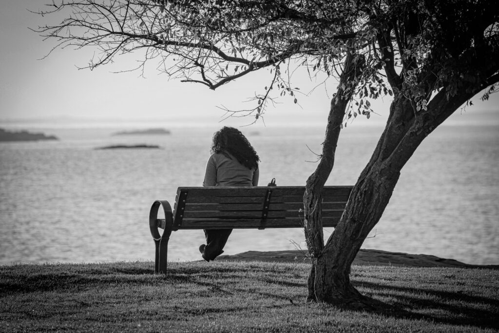 A person sits on a bench, enjoying a tranquil view of the water in black and white.