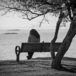A person sits on a bench, enjoying a tranquil view of the water in black and white.