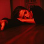 A pensive woman rests her head on a table, bathed in red lighting, reflecting emotion.